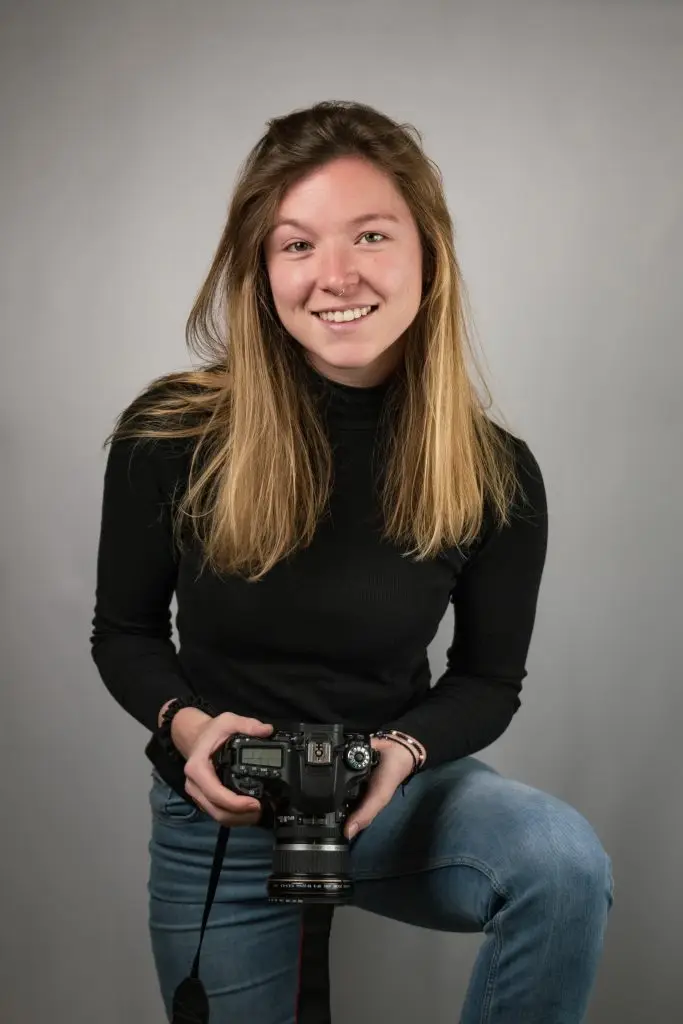 blonde Frau mit schwarzen Rollkragenpulli hält eine Kamera und lächelt