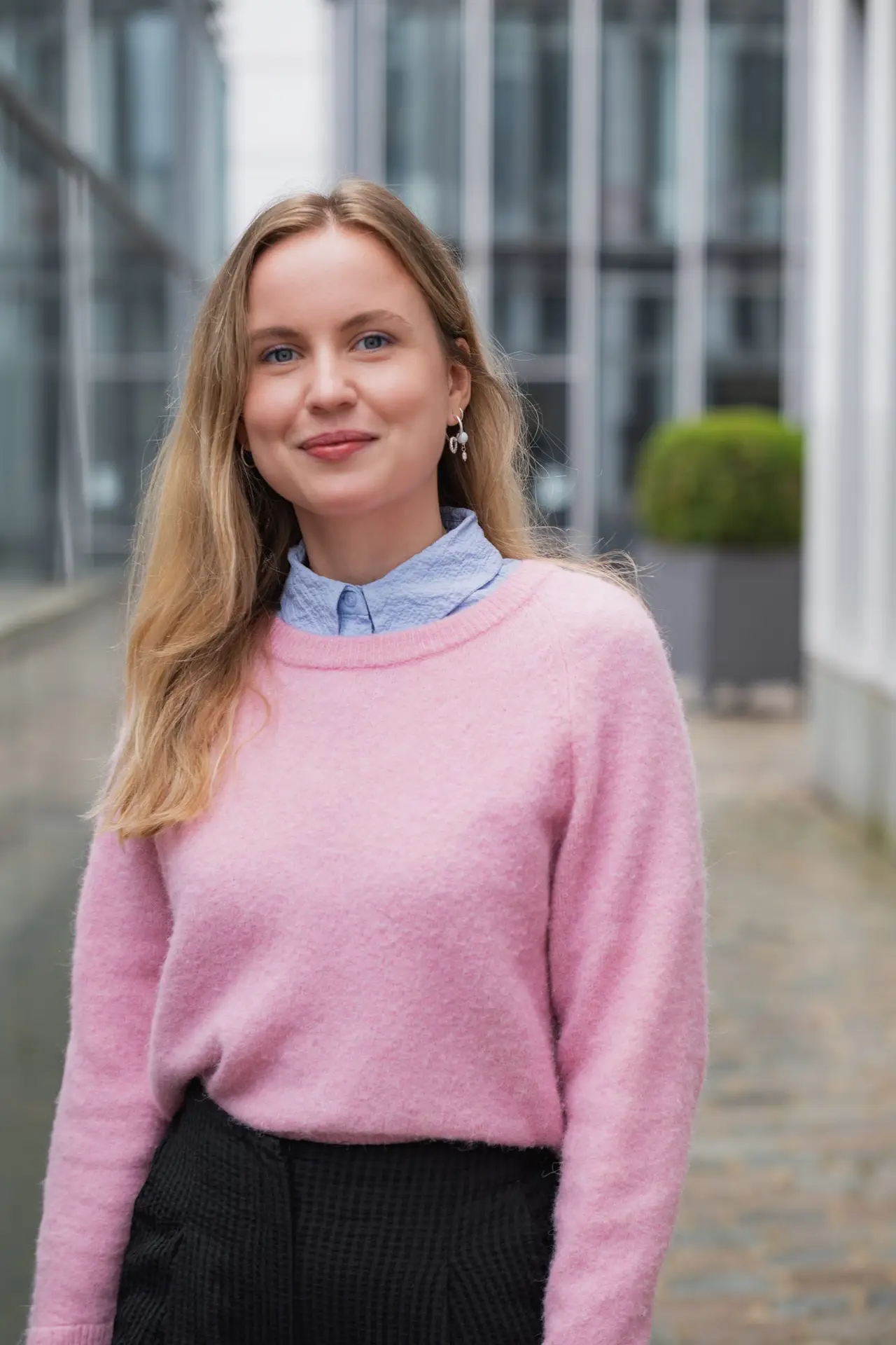 Eine blonde junge Frau mit einem Rosinen Pullover blickt freundlich und zuversichtlich in die Kamera