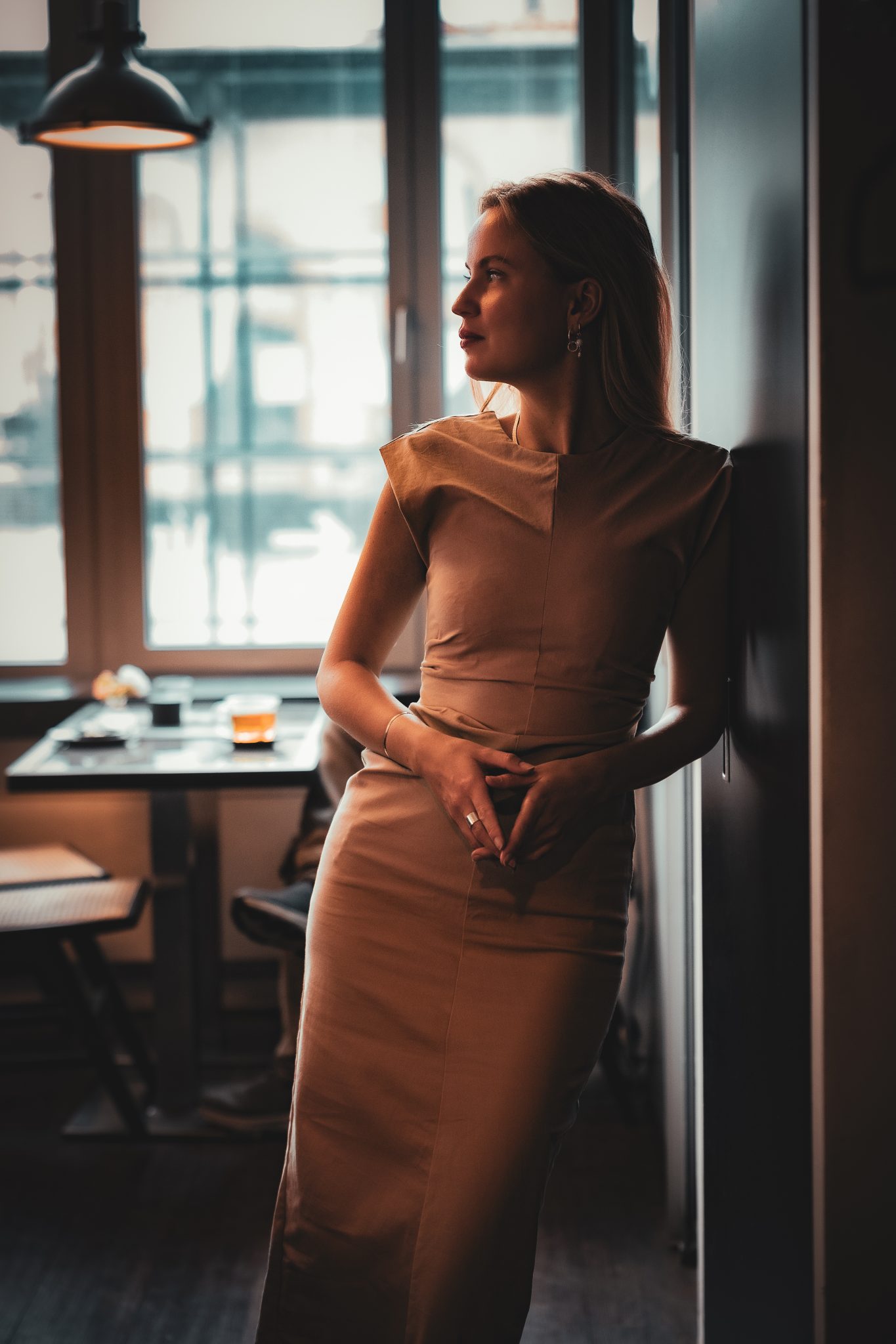 Eine blonde junge Frau steht an eine Wand in einem Café gelehnt und schaut zur Seite. Ihre Hände bilden vor ihrem Körper eine Art Raute