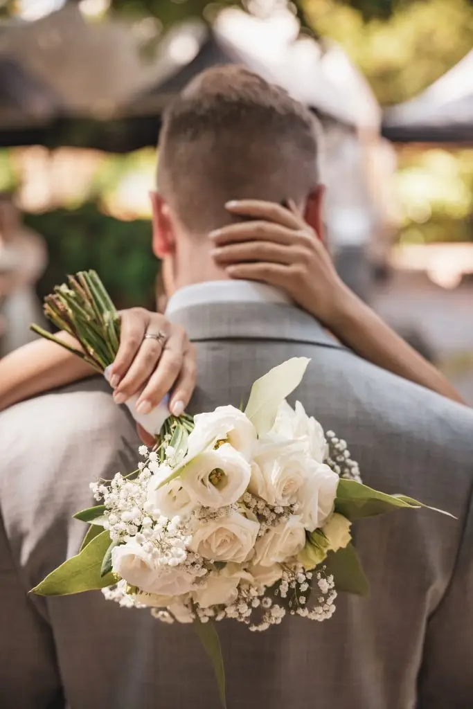 Ein Bräutigam von hinten. Die Braut lässt mit einer Hand an seinen Hinterkopf und hält in der anderen Hand ihren Brautstrauß