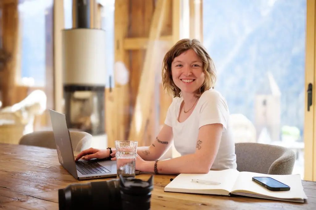 eine blonde Frau sitzt mit ihrem Laptop an einem Tisch. Im Hintergrund ist ein Kamin und Holzwände.