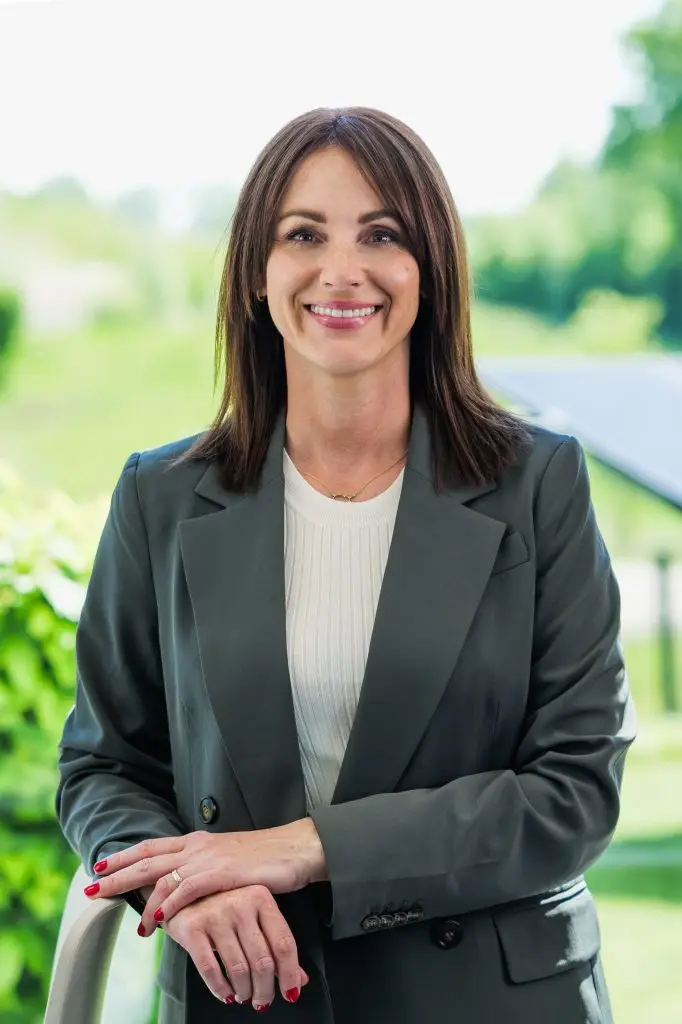 Eine Businessfrau lächelt in die Kamera, im Hintergrund ist ist eine grüne Landschaft und eine Solaranlage