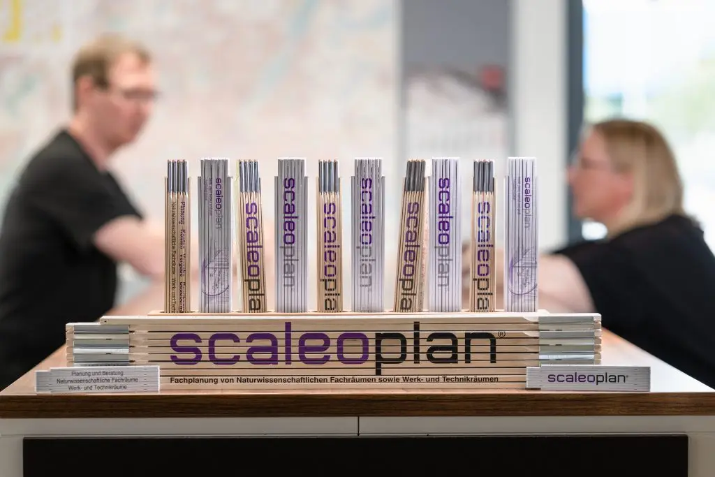 Ein riesiger Zollstock und viele kleine, die senkrecht nach oben zeigen mit dem Schriftzug "scaleoplan" darauf. Dahinter zwei unscharfe Personen, die sich angucken